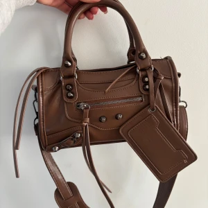Brun handväska i skinn med detaljer - Snygg brun handväska i skinn med silverfärgade nitar, dragkedja framtill och långa dekorativa remmar. Väskan har både handtag och axelrem samt en matchande tag. Perfekt för dig som gillar coola detaljer och stilren design.