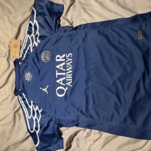 PSG x Jordan blå fotbollströja - Blå PSG x Jordan fotbollströja med vita detaljer och vingar på axlarna. Kortärmad modell i lätt och ventilerande material, perfekt för match eller träning. Stora loggor på bröstet och sponsortryck framtill. Snygg och sportig design för dig som älskar fotboll.