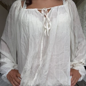 Vit blus i linne med snörning - Luftig och romantisk vit blus i linne med långa ärmar och snörning framtill vid halsen. Blusen har en avslappnad passform och elastiska muddar vid ärmslut och nederkant. Perfekt för en bohemisk look eller varma dagar.