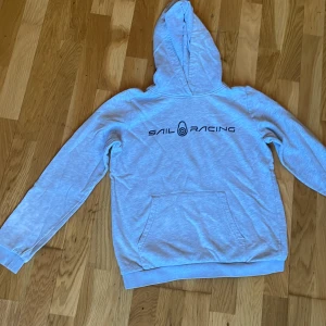 Ljusgrå Sail Racing hoodie med tryck - Ljusgrå hoodie från Sail Racing med svart logotyptryck på ryggen och en stor huva. Klassisk känguruficka framtill och mjukt material som känns skönt mot huden. Perfekt för dig som gillar sportig och avslappnad stil.