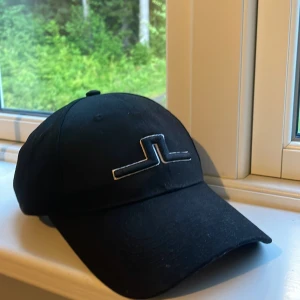 Svart keps från J.Lindeberg - Snygg svart keps från J.Lindeberg med deras ikoniska broderade logga framtill och baktill. Justerbar rem med metallspänne baktill för perfekt passform. Klassisk böjd skärm och ventilerande hål upptill. Perfekt accessoar för en clean och sportig look.