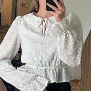 Vit blus - Supersöt vit blus med volangkant nertill, puffiga långärmar och stor krage med knytrosett framtill. Blusen har en lätt skrynklig struktur och ger en romantisk vibe. Perfekt att styla med jeans eller kjol för en trendig look.