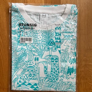 STUNSIG t-shirt från IKEA x Harrington - Unik t-shirt från IKEA STUNSIG-kollektionen, designad av Steven Harrington. Vit bas med turkost grafiskt mönster och coola illustrationer över hela tröjan. Klassisk passform med rund hals och korta ärmar. Tillverkad i mjuk bomull – perfekt för dig som gillar statement-plagg.
