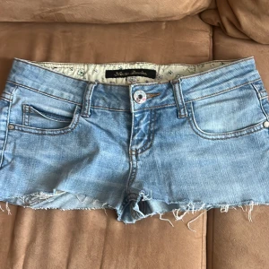 Ljusblå jeansshorts med fransig kant - Snygga ljusblå jeansshorts med fransig, rå kant och klassiska fickor fram och bak. Shortsen har normal passform och low waist, perfekta för sommardagar. Tidlös denimlook med slitna detaljer för en avslappnad vibe. PRIS KAN DISKUTERAS 