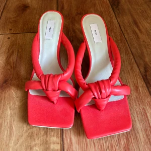 Röda klackade sandaler från Nelly - Snygga röda sandaler från Nelly med fyrkantig tå och knutdetalj framtill. Skorna har vadderade remmar och en låg klack som ger en trendig look. Perfekta för att lyfta din outfit med en pop av färg.