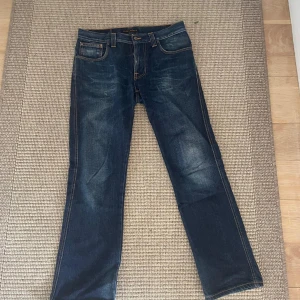 Mörkblå jeans från Nudie Jeans, W32 L30 - Säljer ett par mörkblå jeans från Nudie Jeans med klassisk femficksdesign. Jeansen har raka ben, orangea sömmar och Nudie-logga på bakfickorna. 