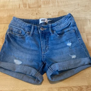 Blå jeansshorts från 157 - Snygga blå jeansshorts från 157 med upprullade benslut och slitna detaljer framtill. Klassisk femficksmodell med knapp och dragkedja. Perfekta för varma dagar och en avslappnad look.