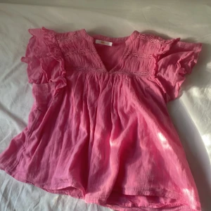 Rosa blus med volang från Gina Tricot - Supersöt rosa blus från Gina Tricot med volangiga korta ärmar och v-ringning. Blusen har smockdetaljer upptill och är tillverkad i ett lätt, luftigt tyg som passar perfekt till varma dagar. Den har en lös och flowy passform som ger en chill vibe. Lappen där det står Gina är lite lös annars är det inget fel på den