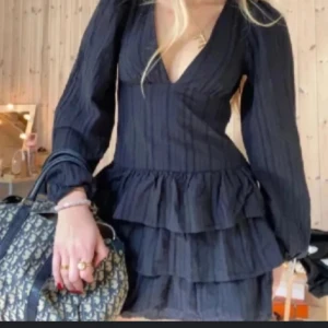 Svart volangklänning med v-ringning - Supersnygg svart kortklänning med djup v-ringning och långa ärmar. Klänningen har flera lager volanger i kjolen och ett diskret randigt mönster i tyget. 