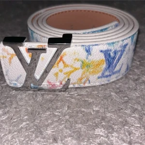 Louis Vuitton bälte färgglatt  - Två snygga Louis Vuitton-bälten med ikoniska LV-spännen i silver. Ena bältet är vitt med färgglada LV-loggor i blått, gult och orange, det andra har ett klassiskt beige och vitt rutmönster. Båda är i präglat skinn och har exklusiv känsla.