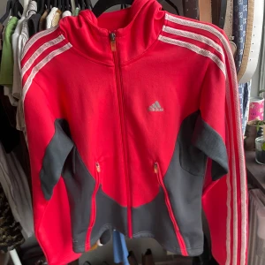 Röd Adidas Huvtröja - Sportig röd och grå hoodie från Adidas med huva, hel dragkedja och klassiska vita ränder längs ärmarna. Jackan har två fickor med dragkedja och Adidas-logga på bröstet. Perfekt för träning.