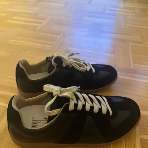 Maison Margiela svarta  - Svarta sneakers från Maison Margiela med beige sula och vita snören. Skorna har en klassisk rund tå, ovandel i skinn och mocka samt diskreta sömdetaljer baktill. Snygg kontrast mellan material och färger, perfekt för dig som gillar stilrena men unika sneakers.