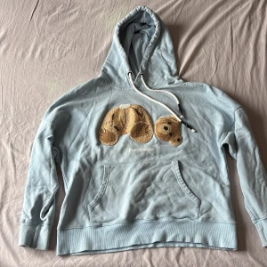 Ljusblå Palm Angels hoodie med björn - Säljer en ljusblå hoodie från Palm Angels med broderad björn på bröstet och vit snörning i huvan. Tröjan har känguruficka och ribbade muddar. Perfekt statement-plagg för dig som gillar streetwear och unika detaljer.