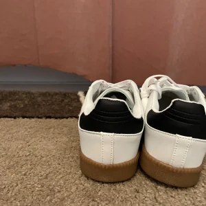 Adidas Samba OG vit/svart/gum - Adidas Samba OG sneakers i vitt skinn med svarta detaljer och klassiska tre ränder. Grå mocka framtill och brun gummisula ger en retro vibe. Guldtexten 'SAMBA' på sidan och blå etikett på plösen. Perfekta för dig som gillar streetstyle.