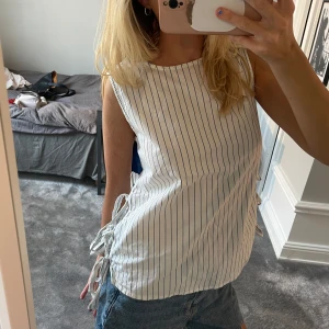 Randig ärmlös linnetopp med knyt - Snygg ärmlös linnetopp i vitt med tunna blå ränder och knytband på båda sidorna. Toppen har rak passform och rund halsringning, perfekt för en sommardag. Tillverkad i lätt bomullsmaterial som känns fräscht och luftigt.