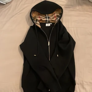 Svart Burberry hoodie med dragkedja - Svart hoodie från Burberry med klassiskt rutigt foder i huvan. Tröjan har dragkedja framtill, snörning i huvan och ribbade muddar. Otroligt skön tröja. OBS! Det står storlek S men skulle säga att den är rätt liten för att vara S. Jag skulle säga att den är XS. 