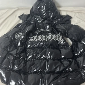 Svart glansig pufferjacka. Fick den som present men jag hade många jackor så försöker sälja den - Svart glansig pufferjacka från Zonerboyz med avtagbara ärmar så du kan bära den som väst. Stor broderad logga på ryggen och mindre logga på bröstet och ärmarna. Jackan har huva, dragkedja och en riktigt fet streetkänsla.