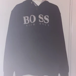 Svart hoodie från Hugo Boss XXL - Svart hoodie från Hugo Boss i storlek XXL med stor BOSS-logga broderad på bröstet. Tröjan har huva, dragkedja framtill och är långärmad. Perfekt för dig som gillar en clean och klassisk streetstil.