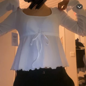 Vit blus med knyt och volang - Supersöt vit blus med långa ärmar, knyt i midjan och volangkant nertill. Blusen har en rundad urringning och är lätt utsvängd nertil.