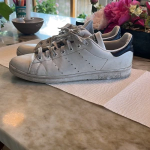Adidas Stan Smith 44,5 - Klassiska Adidas Stan Smith sneakers i vitt skinn med mörkblå detalj på hälen. Perfekt för dig som gillar en clean och tidlös look. Säljer skorna då de är för små. Priset är inte hugget i sten. Hör av dig vid minsta fundering :)