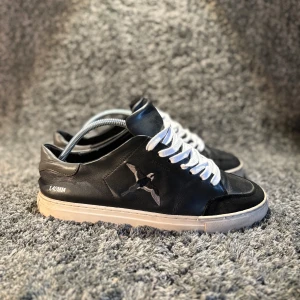 Axel arigatos  - Svarta sneakers i skinn och mocka med vit sula och vita snören. Sidan pryds av ett broderat fågelmotiv. Klassisk låg modell med rund tå och platt sula, perfekt för en stilren och avslappnad look.🌟storlek 42 skick bra 