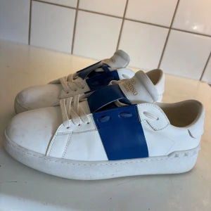 Ljusblå valentino opens - Snygga Valentino Garavani sneakers i vitt skinn med bred blå läderdetalj över snörningen. Klassisk rund tå och platt sula med diskret nitar baktill. Kommer med originalkartong och dustbag. Perfekta för dig som vill sticka ut med stil.