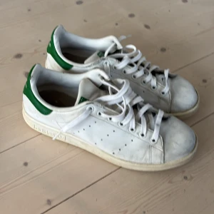 Adidas Stan Smith vita sneakers 40 2/3 - Klassiska Adidas Stan Smith sneakers i vitt skinn med gröna detaljer på hälen och tungan. Skorna har rund tå, platt sula och snörning. 