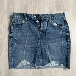 Blå jeanskjol från ONLY med slitningar - Snygg blå jeanskjol från ONLY med rå kant nertill och slitna detaljer framtill. Kjolen har klassisk femficksdesign, synliga knappar och en cool, avslappnad vibe. Perfekt för dig som gillar en trendig och avslappnad stil.