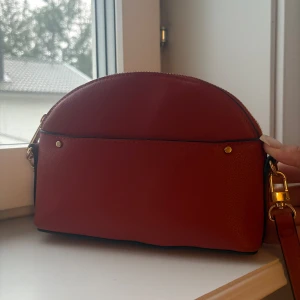 Röd axelväska i skinn med gulddetaljer - Säljer en stilren röd axelväska i skinn med halvmåneform och justerbar axelrem. Väskan har guldfärgade metalldetaljer och dragkedja upptill. Helt ny.