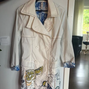 Vit trenchcoat från Desigual med print - Unik vit trenchcoat från Desigual med coola handritade prints i färg nertill och på insidan av ärmarna. Jackan har dubbelknäppning, bred krage och långa ärmar. Köpte för 900 från butik.