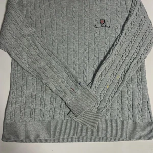 Grå kabelstickad tröja från Barbour - Kabelstickad grå tröja från Barbour med broderad logga på bröstet. Tröjan har rund hals, ribbade muddar och lång ärm. Perfekt för lager-på-lager och chill höst- eller vinterstil. Har lite färg på ärmarna men kan tvättas bort med noggran tvättning
