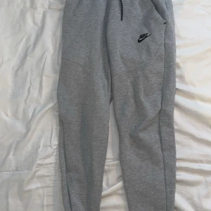 Grå mjukisbyxor från Nike  - Snygga grå mjukisbyxor från Nike med svart logga på ena benet. Byxorna har dragsko i midjan och är tillverkade i mjukt bomullsmaterial. Perfekta för chill eller träning, med en skön loose passform och praktiska fickor. Storleken är XS i herr storlek. Och XL i kids