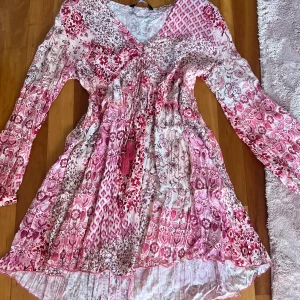 Rosa mönstrig klänning  - Supersöt kortklänning från Zara i rosa och vitt med blommigt mönster. Klänningen har långa ärmar, v-ringning och snörning framtill med tofsar. Luftigt material, perfekt för varma dagar. Fina volangdetaljer! Aldrig använt. 