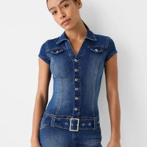 Kort jeansklänning med bälte - Snygg kort jeansklänning i blå denim med knappar framtill, krage och korta ärmar. Klänningen har markerad midja, bälte med spänne och fickdetaljer på bröstet. Använd 1 gång.