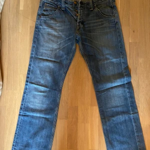 Blå raka jeans från Lee W34 L32 - Säljer ett par klassiska blå jeans från Lee i modellen Flint 3 Needle. Jeansen har raka ben, normal passform och snygga slitningar framtill. Tillverkade i bomull med Lee-logga på bakfickan och läderpatch i midjan. Perfekta för en avslappnad stil.