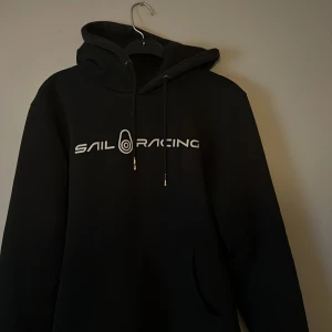 Svart Sail Racing hoodie med tryck - Svart hoodie från Sail Racing med vit logga framtill och tryck på huvan. Klassisk modell med dragsko i huvan och känguruficka. Perfekt för dig som gillar stilren streetwear och vill ha något bekvämt och snyggt.