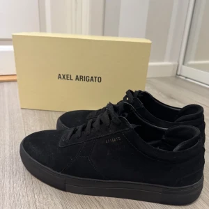 Svarta sneakers från Axel Arigato mocka - Stilrena svarta sneakers från Axel Arigato i mjuk mocka med diskret guldfärgad logga på sidan. Klassisk låg modell med snörning och platt sula. Perfekta för dig som gillar minimalistisk och clean stil. Ny pris 1950kr o Tveka inte att höra av dig vid intresse.