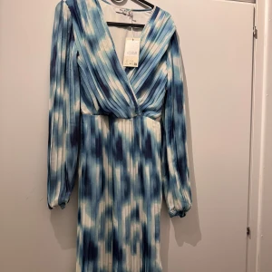 Plisserad blåvit omlottklänning NA-KD - Superfin långklänning från NA-KD med plisserat tyg och omlott-v-ringning. Klänningen har ett blåvitt mönster, långa ärmar med resår vid handlederna och en flowy siluett. Perfekt för dig som vill sticka ut med en cool och fräsch look.