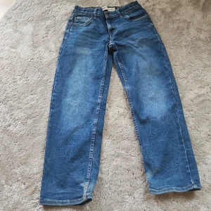 Levi's 501 raka blå jeans - Klassiska Levi's 501 jeans i blå denim med raka ben och normal passform. Jeansen har fem fickor, synliga sömmar och den ikoniska röda Levi's-taggen på bakfickan. Perfekta för en avslappnad och tidlös stil.