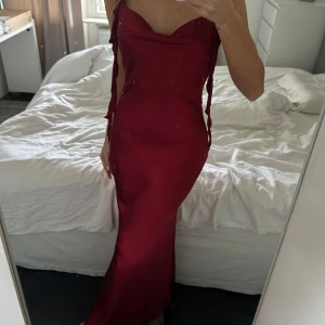Vinröd långklänning med snörning - Säljer en vinröd långklänning i satinliknande material med smickrande figurnära passform. Klänningen har smala axelband med snörningsdetaljer och öppen rygg. Perfekt för dig som vill sticka ut med en elegant och trendig look.