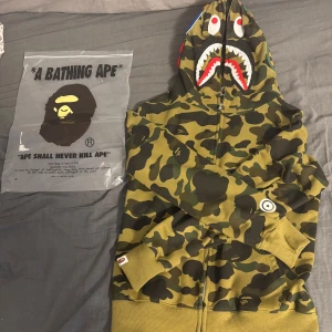 BAPE camo zip hoodie med shark-hood - Säljer en ikonisk BAPE camo zip hoodie med shark-motiv på huvan och broderade bokstäver. Klassisk grön camouflage, dragkedja hela vägen och feta detaljer som tiger- och hajansikte. Perfekt för dig som vill sticka ut med streetstyle.