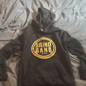 Svart Grind Gang hoodie med guldtryck - Svart hoodie från Grind Gang Music med stort guldtryck på bröstet. Klassisk känguruficka framtill och huva med snörning. Perfekt för dig som gillar streetwear och vill sticka ut med en fet logga.