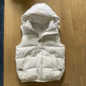 Vit dunväst med huva från Zara - Snygg vit dunväst från Zara med stor huva och quiltad design. Västen har två stora fickor framtill med dragkedjedetaljer och stängs med både dragkedja och tryckknappar. Perfekt för lager-på-lager och chill stil.