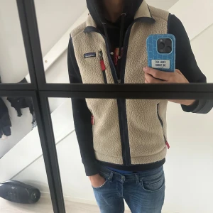 Beige fleeceväst från Patagonia - Snygg beige fleeceväst från Patagonia med svarta detaljer och röda dragkedjor. Västen har hög krage, tre fickor med dragkedja och klassisk Patagonia-logga på bröstet. Perfekt lager-på-lager-plagg för kyliga dagar.