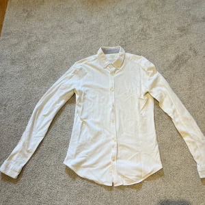 Vit slim fit skjorta från Zara Man - Vit skjorta från Zara Man i slim fit-modell med klassisk krage och knäppning framtill. Skjortan har långa ärmar med ljusgrå armbågslappar. Den är i storlek M