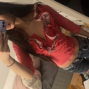 Röd blus med rutiga volanger - Säljer en röd blus med trekvartsärm. Blusen har volanger längs urringningen och framtill, med inslag av rödvitt rutigt tyg som ger en unik look. Knappar framtill och figurnära passform. Perfekt för dig som vill sticka ut!