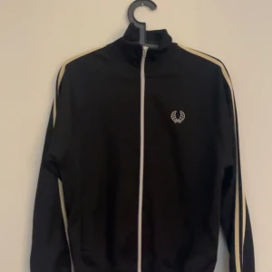 Svart Fred Perry track jacket XS - Svart track jacket från Fred Perry i storlek XS. Jackan har vita ränder längs ärmarna, dragkedja framtill och klassisk broderad logga på bröstet. Perfekt för en sportig och stilren look.