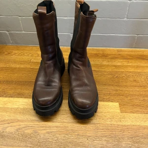Bruna chunky chelsea boots i skinn - Snygga bruna chelsea boots från Tamaris med grov sula och högt skaft. Skorna är i skinn med svarta elastiska paneler på sidorna och dragflikar upptill. Perfekta för dig som gillar en robust och trendig look till höst och vinter. Skorna är en stor strl 39
