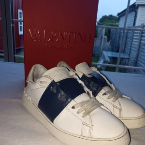 Valentino Garavani Open sneakers vit/blå - Snygga Valentino Garavani Open sneakers i vitt skinn med bred marinblå läderdetalj över mitten. Klassisk rund tå, platt sula och coola nitar baktill. Vit snörning och diskret logga på plösen. Persfekta för dig som gillar lyxig streetstyle. Kvitto finns. De är äkta. Hör av er om ni har några frågor.