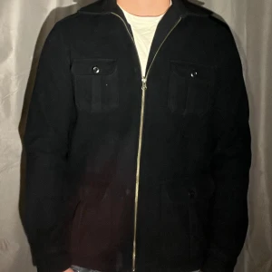 Mörkblå overshirt med dragkedja - Stilren svart overshirt med dragkedja framtill och fyra fickor med lock och knapp. Jackan har klassisk krage och raka ärmar, perfekt för lager-på-lager. Materialet är mjukt och ger en clean look som funkar till många outfits.